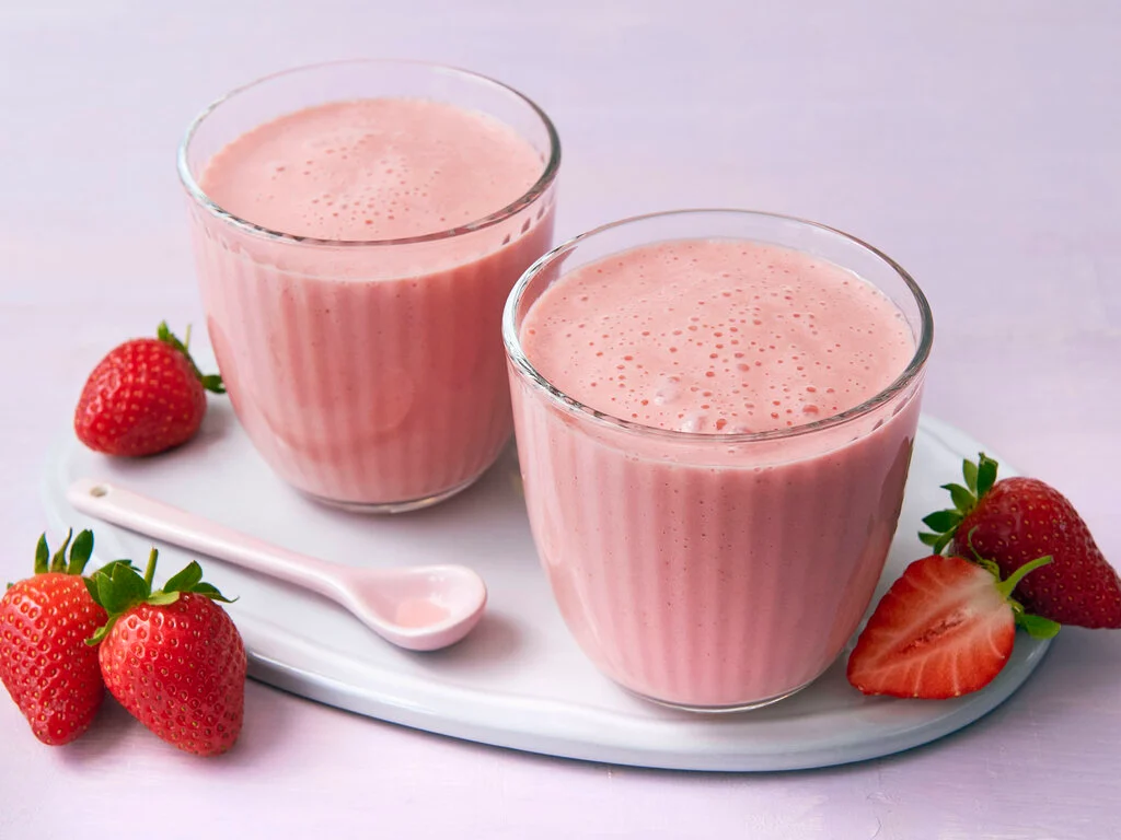 Zwei Gläser mit rosa Erdbeersmoothie auf weißem Tablett, umgeben von frischen ganzen und halbierten Erdbeeren und einem weißen Löffel.