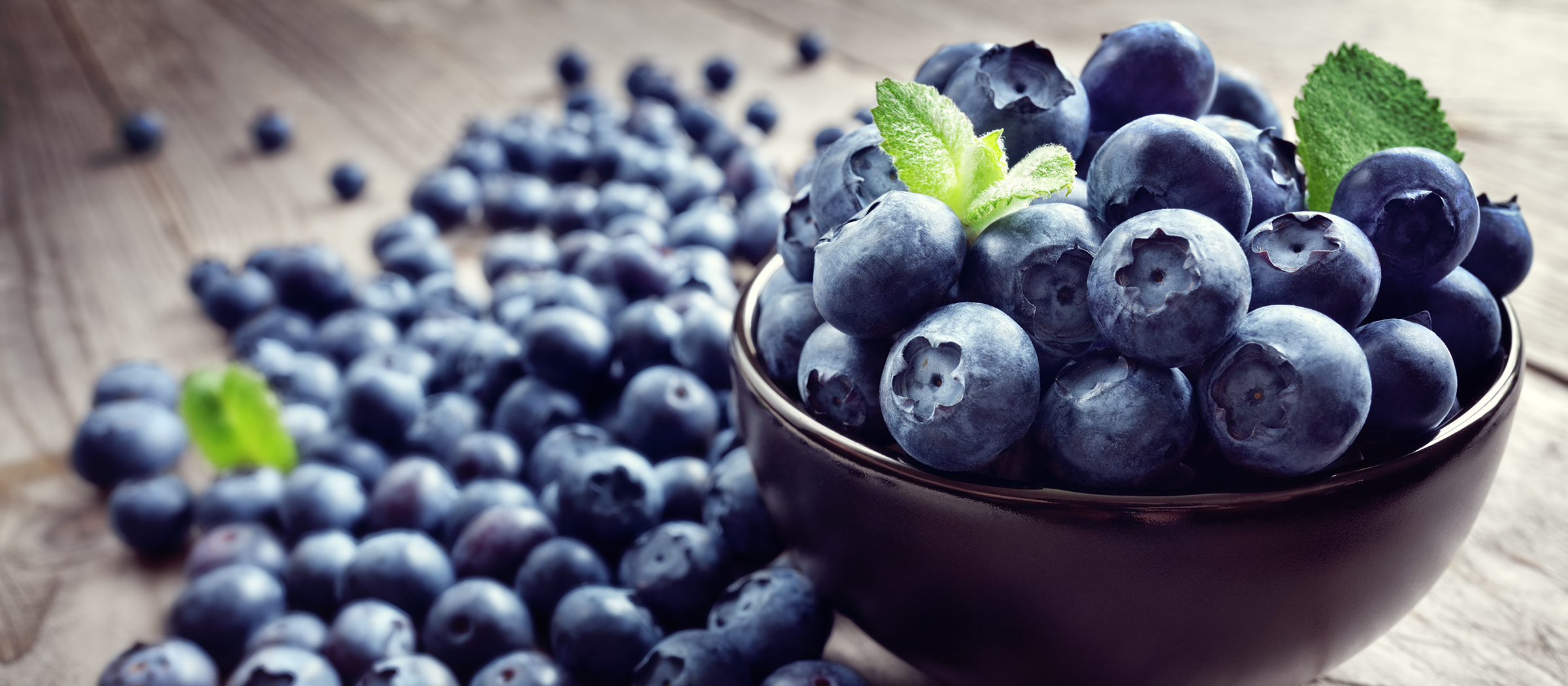 Nahaufnahme einer schwarzen Schale, gefüllt mit frischen Heidelbeeren und einem Minzblatt, daneben weitere Heidelbeeren auf Holzuntergrund.