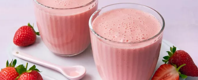 Zwei Gläser mit rosa Erdbeersmoothie auf weißem Tablett, umgeben von frischen ganzen und halbierten Erdbeeren und einem weißen Löffel.