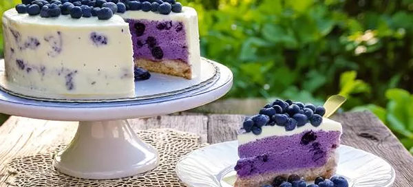 Blaubeer-Joghurt-Torte mit weißer Glasur und Blaubeeren auf einem weißen Tortenständer, ein Stück auf einem weißen Teller mit Blaubeeren daneben.