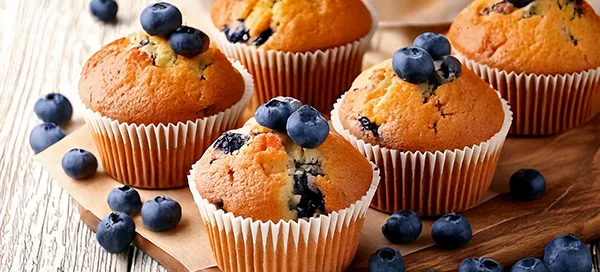 Fünf Blaubeermuffins mit frischen Blaubeeren auf einem Holzbrett, umgeben von verstreuten Blaubeeren.