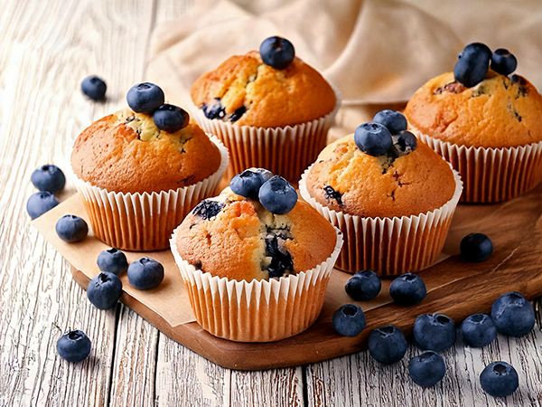 Fünf Blaubeermuffins mit frischen Blaubeeren auf einem Holzbrett, umgeben von verstreuten Blaubeeren.