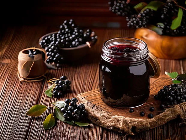 Aroniabeeren-Konfitüre in einem Glas auf einem Holztisch. Aroniabeeren zur Dekoration neben und hinter der Konfitüre in Schalen und auf dem Tisch.