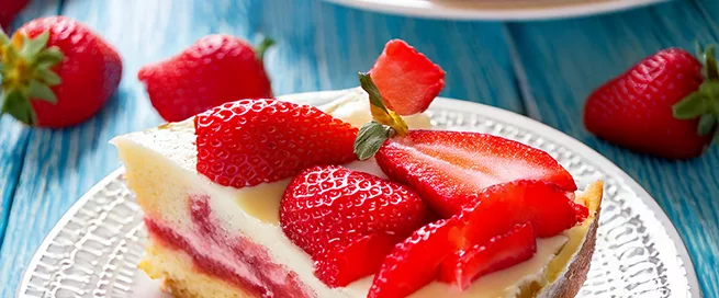 Ein Stück Erdbeerkuchen liegt auf einem weißen, gemusterten Teller. Der Kuchen besteht aus einem hellen Boden, einer Schicht Vanillepudding und frischen, halbierten Erdbeeren als Belag. Im Hintergrund steht ein weiteres Stück auf einem zweiten Teller. Die Oberfläche des Kuchens ist glatt, die Erdbeeren glänzen und sind gleichmäßig verteilt. Der Teller steht auf einer blauen Holzoberfläche. Ein metallener Kuchengabel liegt am Rand des Tellers.