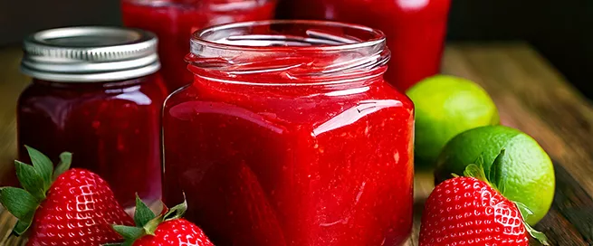 Im Vordergrund steht ein Glas mit leuchtend roter Erdbeermarmelade auf einer Holzoberfläche. Daneben liegen mehrere frische Erdbeeren und zwei Limetten. Im Hintergrund sind weitere Marmeladengläser mit unterschiedlich farbigen Deckeln zu sehen. Die Szene ist hell ausgeleuchtet und zeigt kräftige Farben.