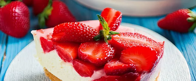 Ein Stück Erdbeerkuchen mit heller Creme und frischen Erdbeeren liegt auf einem weißen Teller. Der Kuchenboden ist goldbraun und wirkt fest. Im Hintergrund steht eine weitere Kuchenplatte mit mehreren Stücken, daneben liegen lose Erdbeeren. Die Oberfläche des Kuchens ist mit halbierten Erdbeeren dicht belegt. Der Tisch ist aus hellblauem Holz.