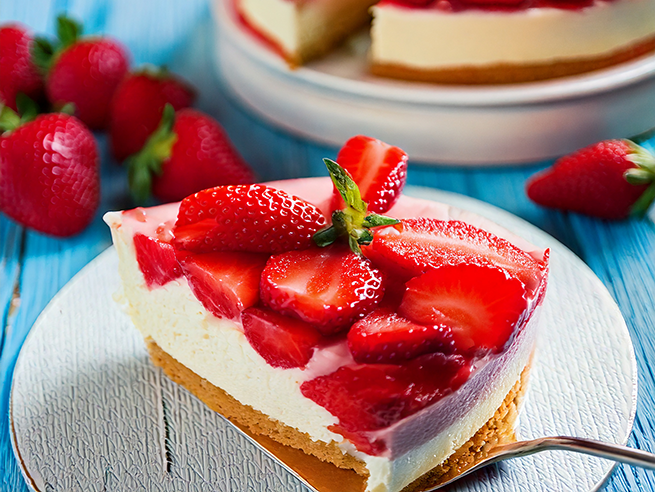 Ein Stück Erdbeerkuchen mit heller Creme und frischen Erdbeeren liegt auf einem weißen Teller. Der Kuchenboden ist goldbraun und wirkt fest. Im Hintergrund steht eine weitere Kuchenplatte mit mehreren Stücken, daneben liegen lose Erdbeeren. Die Oberfläche des Kuchens ist mit halbierten Erdbeeren dicht belegt. Der Tisch ist aus hellblauem Holz.