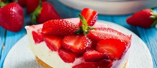 Stück Erdbeerkuchen mit Sahneschicht und frischen Erdbeeren auf einem Kuchenteller, im Hintergrund weitere Erdbeeren und ein angeschnittener Kuchen.