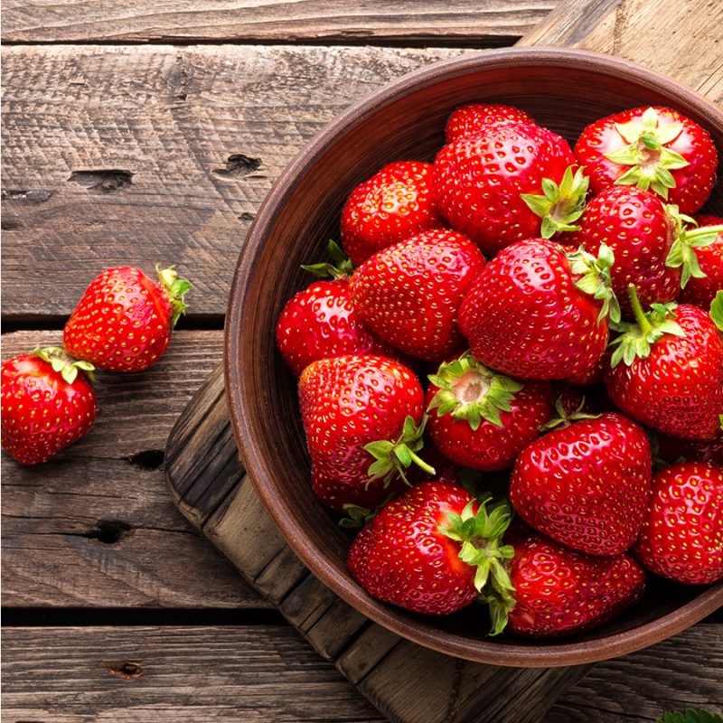 Braune Schale mit mehreren roten Erdbeeren und grünen Blättern auf Holzbrett, daneben einzelne Erdbeeren.