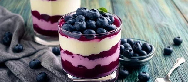 Zwei Gläser mit geschichtetem Heidelbeer-Dessert und frischen Heidelbeeren auf einem Holztisch neben einer kleinen Schale und einem Löffel